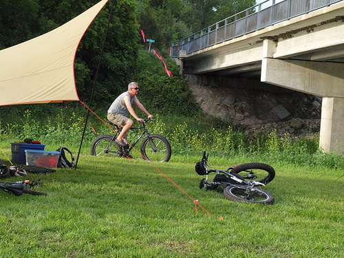Right side view of right of a cyclist riding a black Surly bike, next to a campsite, in a grass field below a bridge